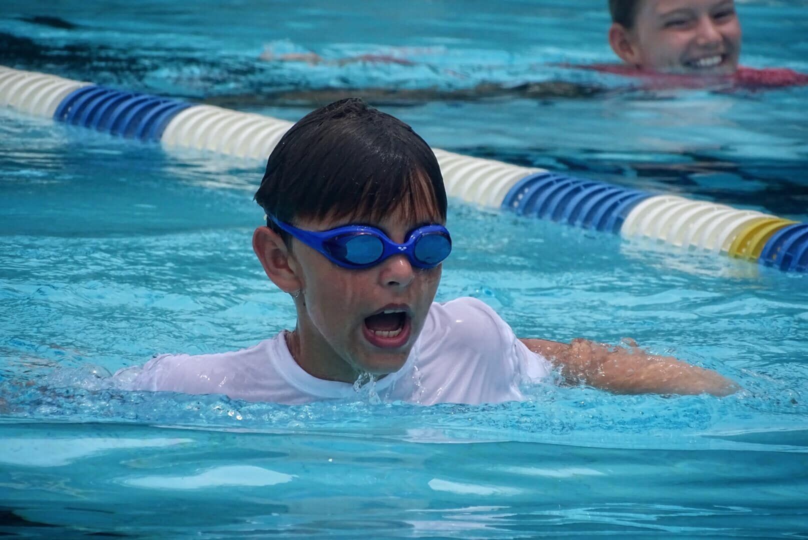 A child wearing blue goggles swims in a pool while another person smiles in the background.