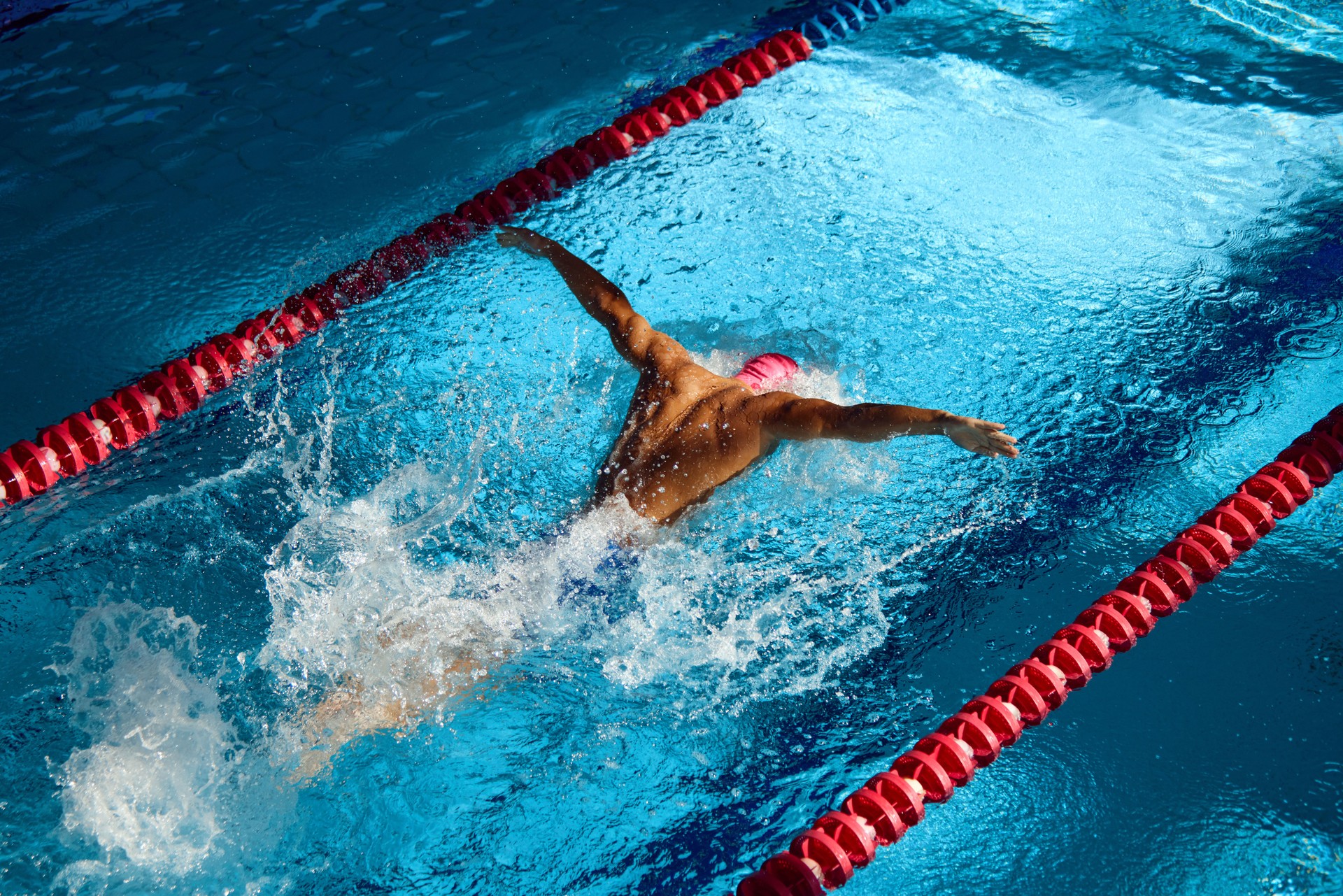 Swimmer in pink cap executes powerful butterfly stroke, his muscular form cutting through blue water, creating dynamic splash between red lane dividers.